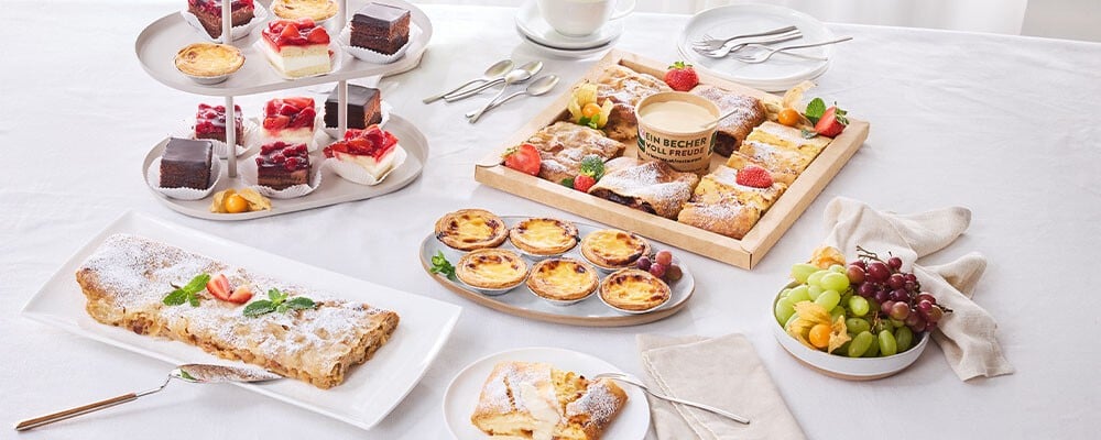 Eine gedeckter Tisch mit verschiedenen Desserts: Apfelstrudel, Pasteis de Nata, Mini-Schnitten