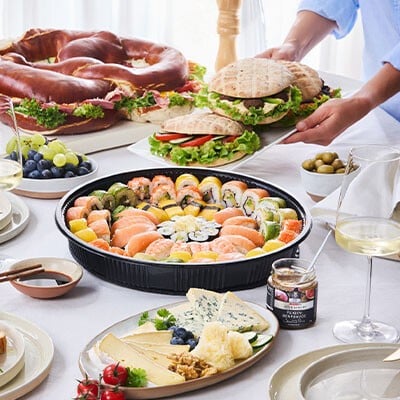 Ein gedeckter Tisch mit verschiedenen Speisen aus dem INTERSPAR Partyservice: Käseplatten, vegetarisch belegte Brötchen, Pita-Brote, Sushi, eine Partybreze und Co