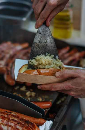 Eine Person legt Sauerkraut mit einem Spachtel auf einen Hotdog 