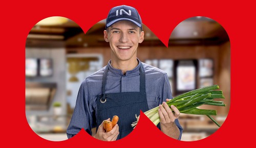 Ein lächelnder junger Mitarbeiter der INTERSPAR-Gastronomie mit einem Lauch und Karotten in den Händen
