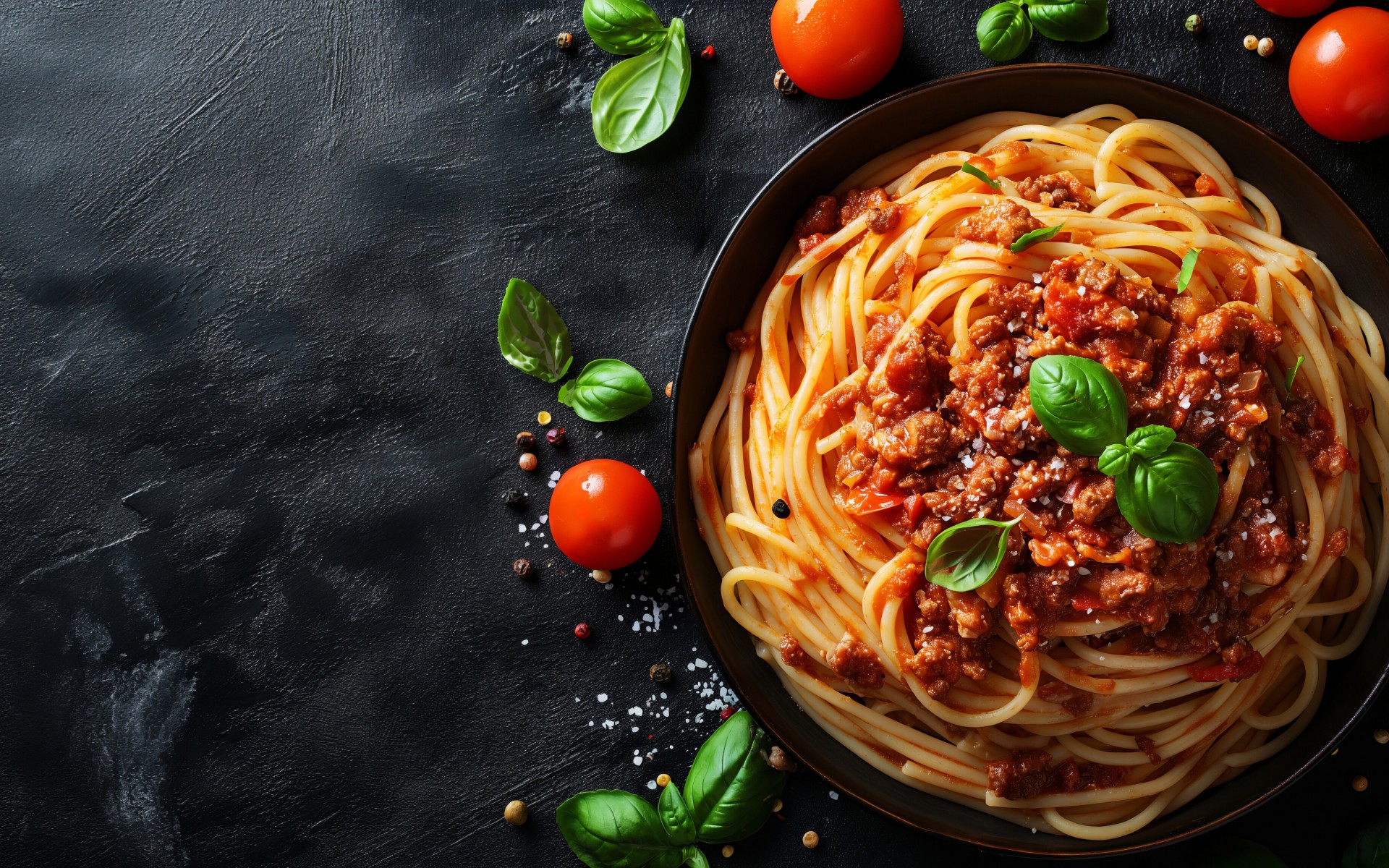 Ein Teller Spaghetti Bolognese neben ein paar Tomaten auf einem dunklen Untergrund