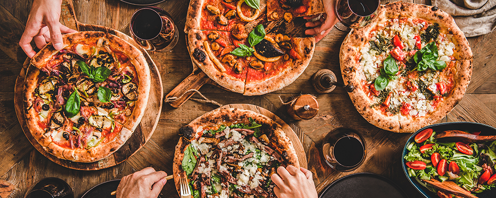 Mehrere verschiedene Pizzen auf einem Tisch an denen sich Leute bedienen - Rezeptbild aus der SPAR Mahlzeit