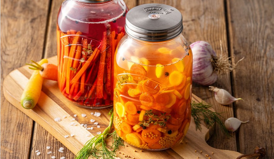 Eingelegte und fermentierte Karotten im Glas, auf einem Holztisch. Rundherum Zutaten und Zubehör zum Einlegen.