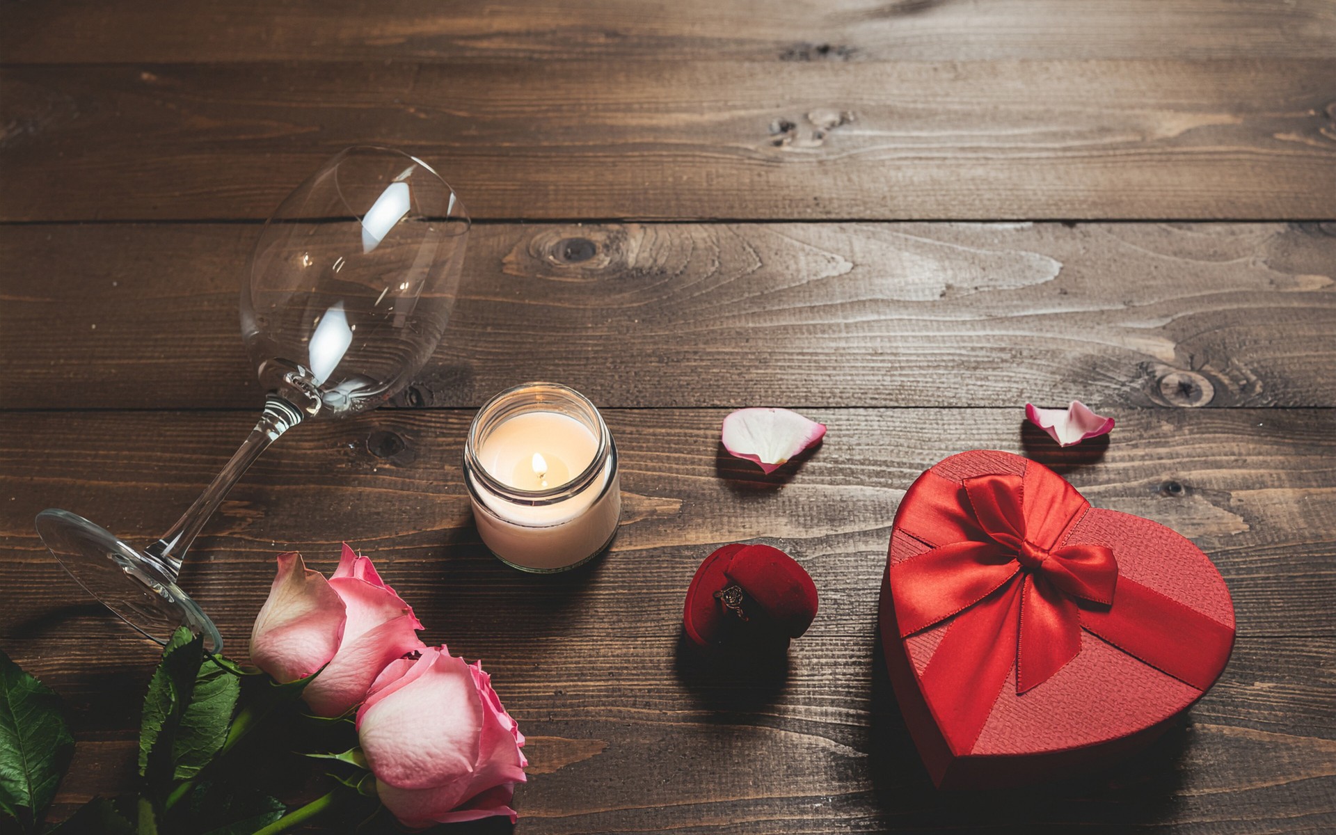 Ein Weinglas, rosa Rosen, ein rotes, herzförmig verpacktes Geschenk und eine Kerze auf einem brauen Tisch