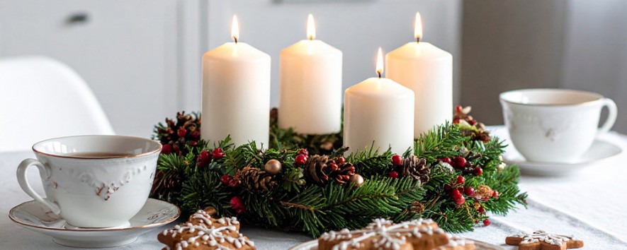 Ein Adventkranz mit vier brennenden Kerzen steht auf einem mit Teetasen und Lebkuchen gedeckten Tisch