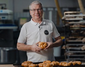 Der/die Backetubenleiter:in lächelt mit seinem/ihrem Bäckerliebling in der Hand in die Kamera