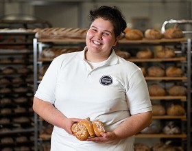 Der/die Backetubenleiter:in lächelt mit seinem/ihrem Bäckerliebling in der Hand in die Kamera