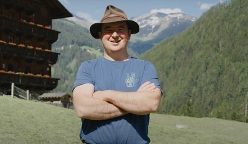 Ein Mann mit einem Hut verschränkt die Arme und lächelt in die Kamera