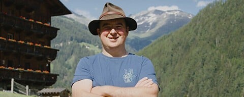 Ein Mann mit einem Hut verschränkt die Arme und lächelt in die Kamera