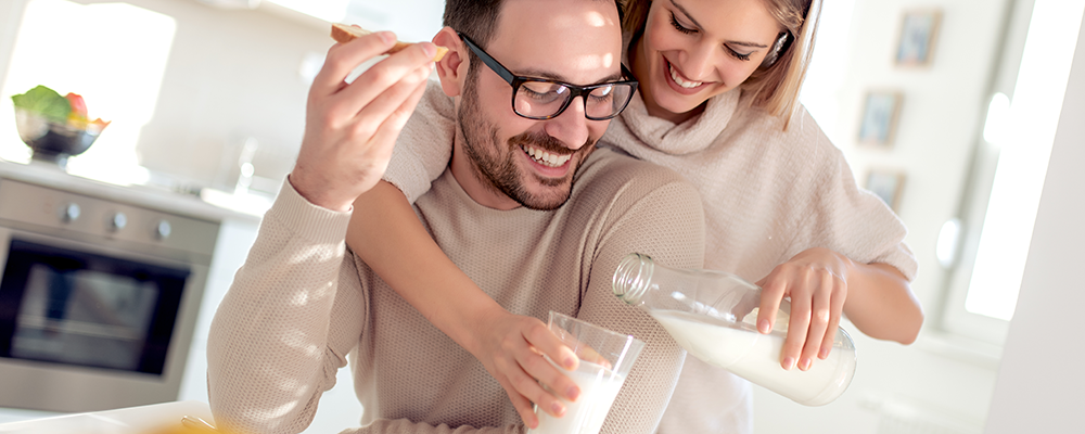 Eine Frau umarmt einen sitzenden Mann von hinten und schenkt ein Glas Milch ein