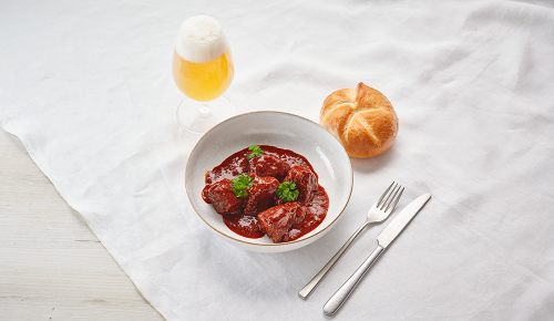 Ein Teller Gulasch mit einer Semmel und einem Glas Bier