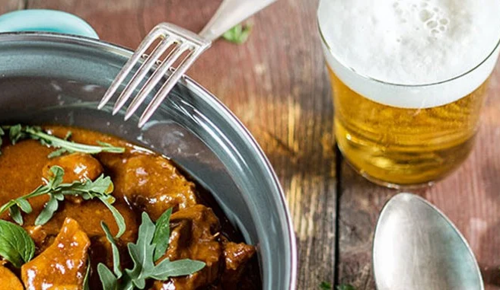 Ein Suppentopf gefüllt mit Gulasch neben einem Glas Bier