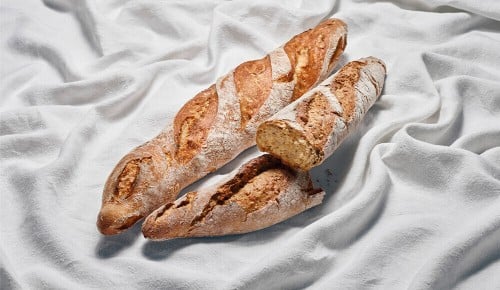 Zwei rustikale Baguettes wovon eines halbiert ist auf einem weißen  Tuch
