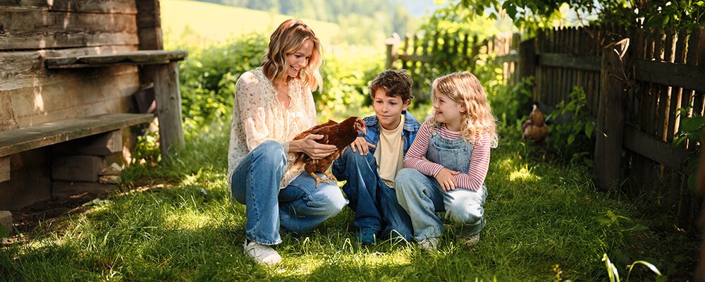 Mirjam Weichselbraun hält ein Huhn in der Hand und hockt neben Kindern in einer grünen Wiese