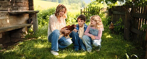 Mirjam Weichselbraun hält ein Huhn in der Hand und hockt neben Kindern in einer grünen Wiese