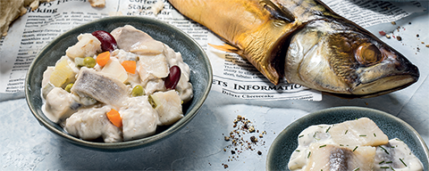 Zwei Schalen mit verschiedenen Fischsalaten und ein geräucherter Fisch auf einer Zeitung