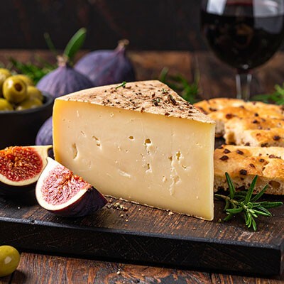 Ein Stück italienischer Asiago auf einem Holzbrett serviert, mit etwas Focaccia, Oliven und Feigen als Beilage