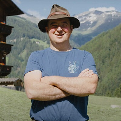 Ein Mann mit einem Hut verschränkt die Arme und lächelt in die Kamera