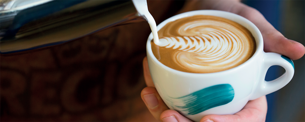 Eine REGIO-Tasse in der eine Person ein Muster mit Milch macht