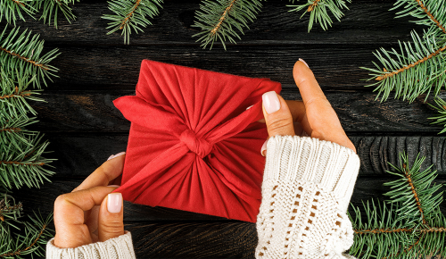 Die Hände einer Frau in einem weißen Häkelpullover, die ein rotes Furoshiki-Tuch über einem Weihnachtsgeschenk bindet