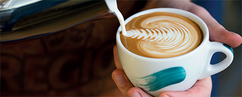 Eine REGIO-Tasse in der eine Person ein Muster mit Milch macht