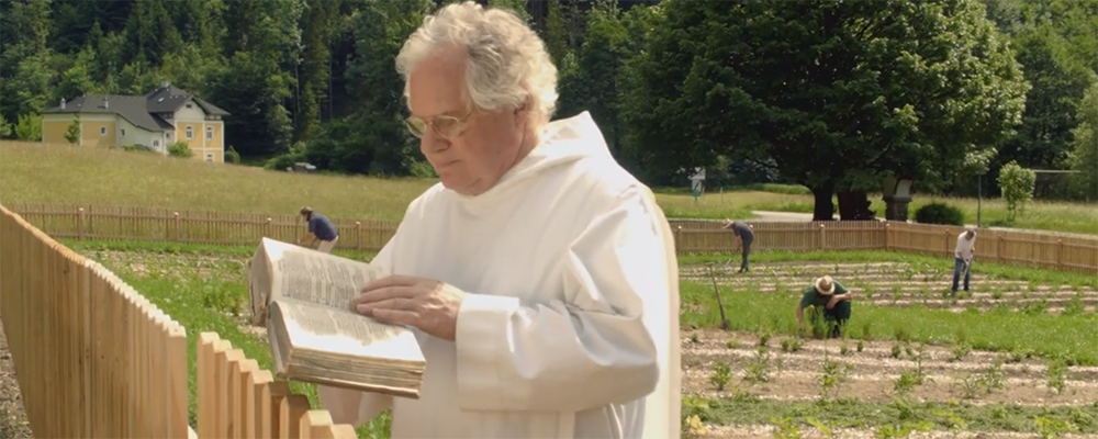 Pater Dr. Johannes Pausch liest in einem alten Buch und steht in seinem Kräutergarten