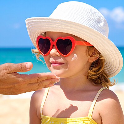 Eine Hand tragt einem Kind mit roter Sonnenbrille und Strohhut - Sonnencreme auf.