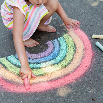 Ein kleines Mädchen malt einen Regenbogen mit Straßenkreide auf Asphalt