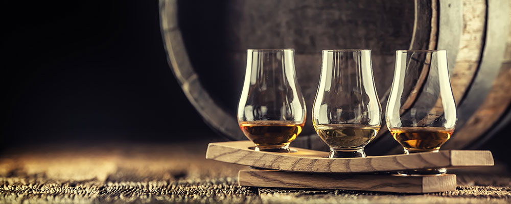 Mehrere Whiskeygläser nebeneinander vor einem Holzfass